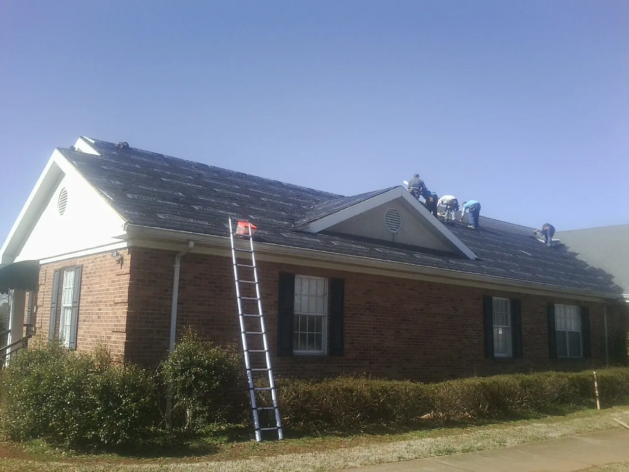 Crew tearing shingles off a brick ranch roof with a ladder set — Emergency Roof Repair work by our crew in Walkertown