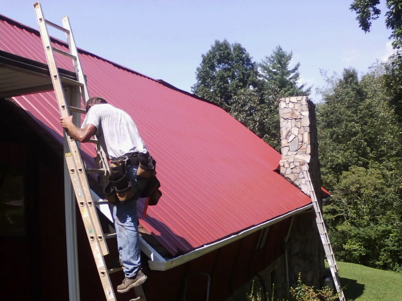 Roofer on a ladder beside a red metal roof and stone chimney — Soffit & Fascia Repair by Roofing Brothers in Walkertown